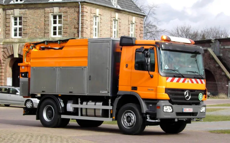 Orange high-pressure jetting vehicle on a Mercedes chassis with stainless steel equipment cabinets in front of a historic brick building