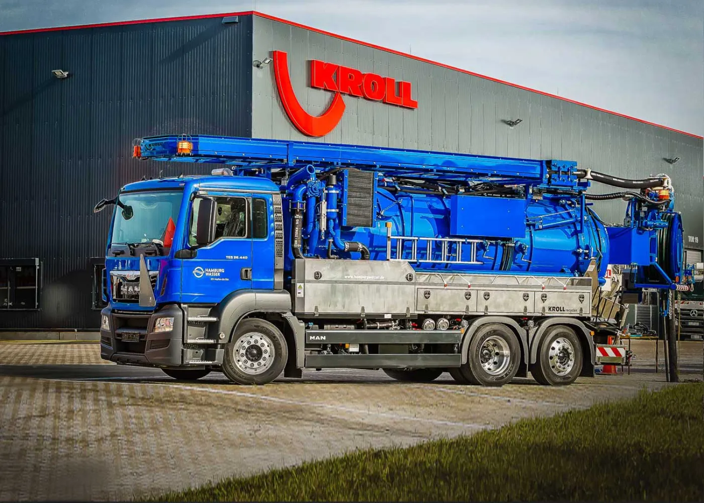 Niebieski pojazd ssąco-płuczący KROLL z długim wysięgnikiem przed halą produkcyjną z napisem KROLL